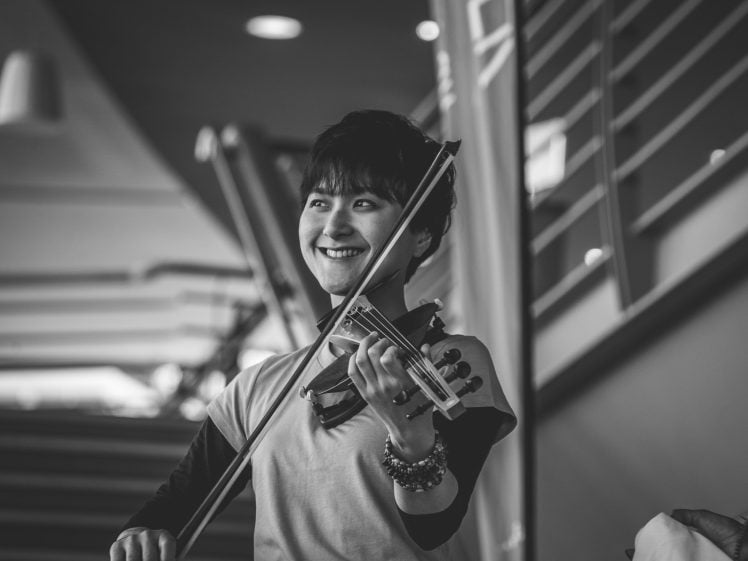 Image of A person smiles whilst playing a violin indoors. The image is in black and white, with a staircase and bannister visible in the background.