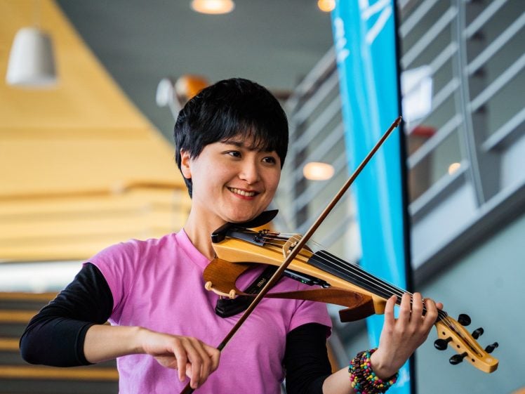Image of A smiling person wearing a pink shirt plays an electric violin indoors, with colourful bracelets on their wrist and a blurred staircase in the background.