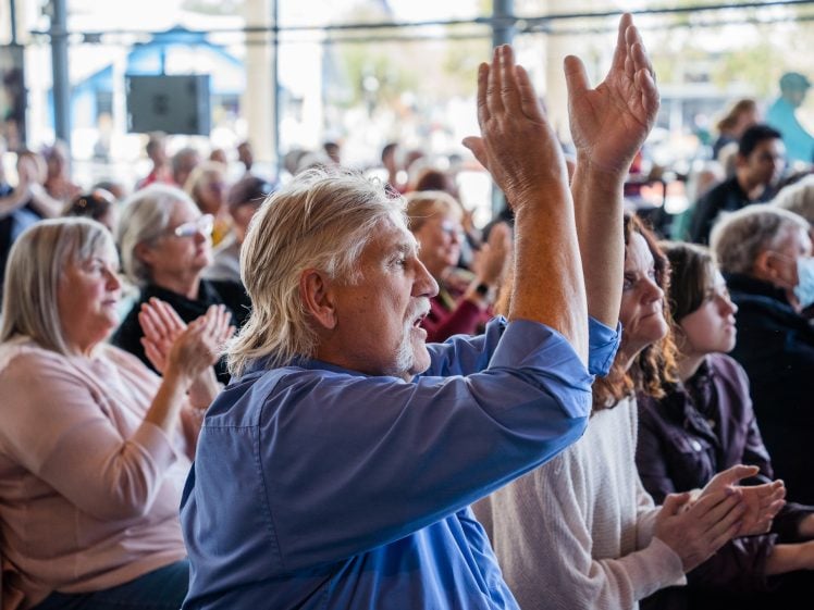 Image of A group of people sit closely together indoors, applauding enthusiastically. The focus is on a man in a blue shirt in the foreground with his hands raised, clapping, surrounded by others who are also clapping and paying attention.