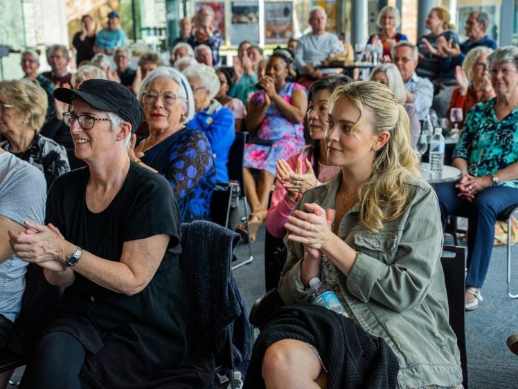 Image of A diverse group of people, mostly older adults, sit closely together in a bright room, smiling and applauding as they watch an unseen performance or speaker.