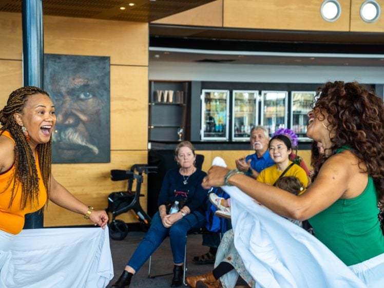 Image of Two women dance and smile energetically in front of an engaged seated audience in a bright, modern room. A large portrait of a man smoking is visible on the wall behind them.