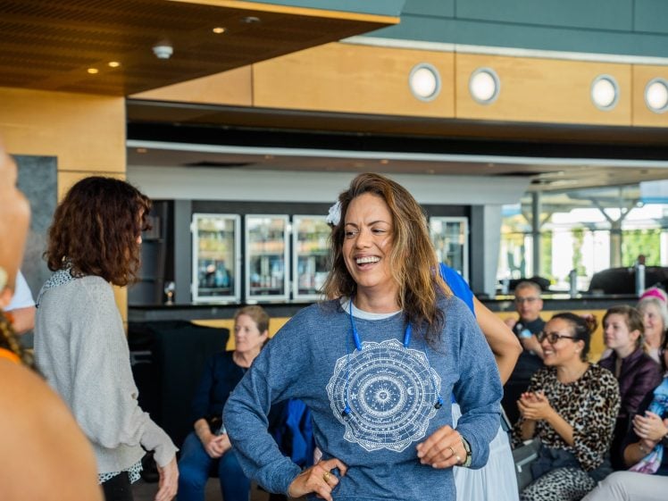 Image of A smiling woman with a flower in her hair stands in the centre of a room, surrounded by people sitting and talking. She wears a blue shirt with a mandala design. The atmosphere appears lively and social.