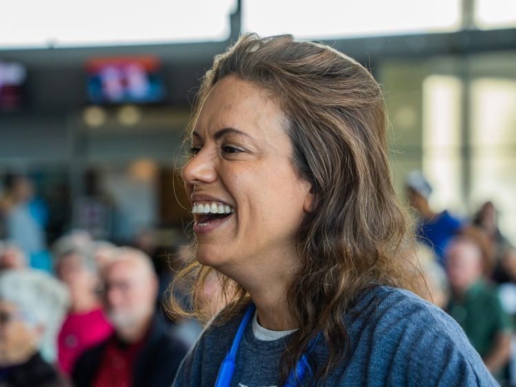 Image of A woman with long brown hair smiles and laughs in a busy indoor setting, with blurred people and screens in the background.