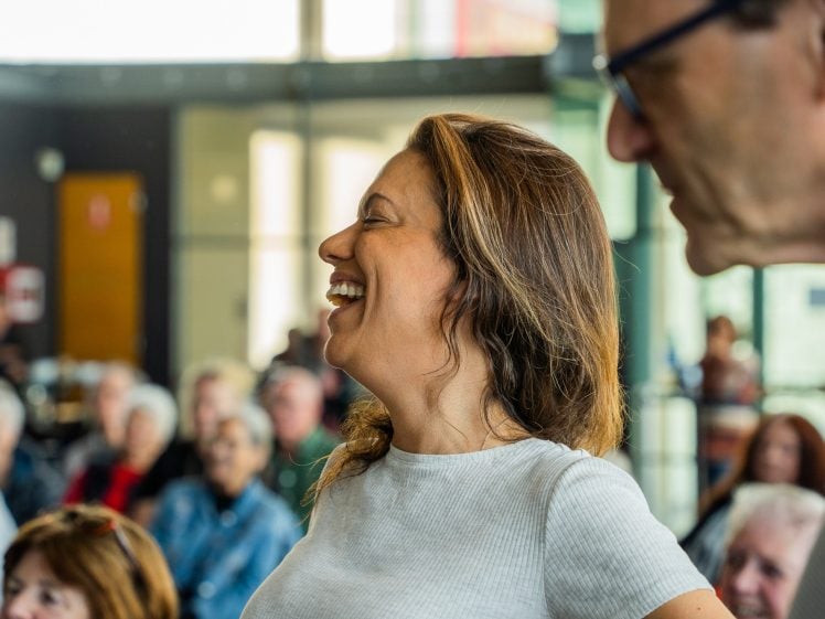 Image of A woman with long brown hair smiles and laughs in a lively indoor setting, with blurred people seated in the background, suggesting a social or community event.
