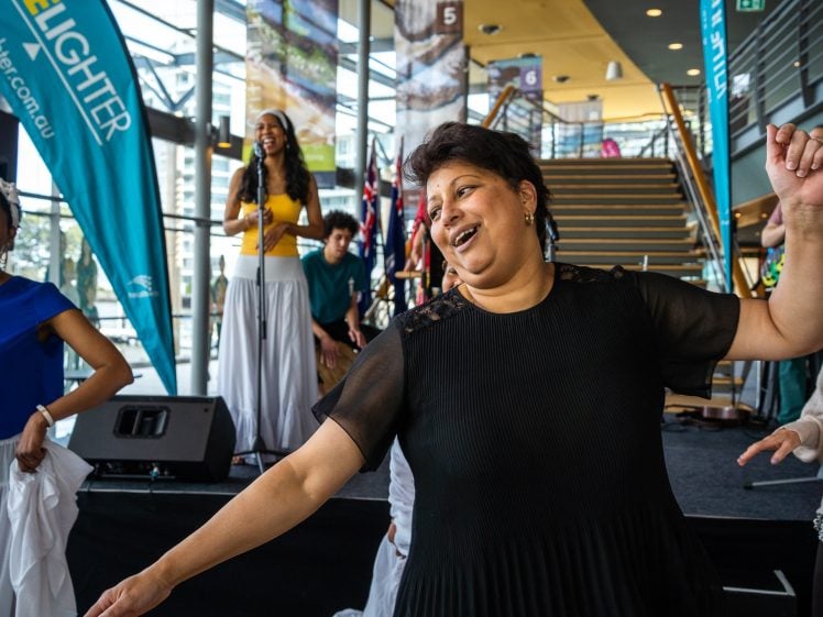 Image of A woman in a black top joyfully dances indoors near a stage, whilst others dance and a woman sings into a microphone in the background. The atmosphere is lively and celebratory.