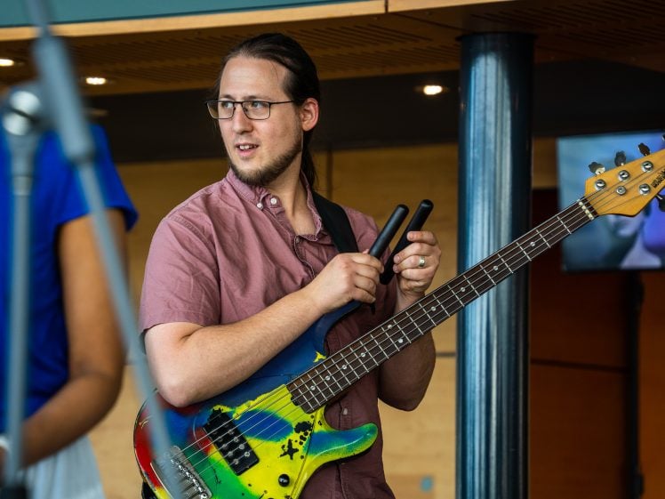 Image of A man with glasses and a brown shirt holds a colourful electric bass guitar and mallets, standing indoors. Another person in a blue shirt is partially visible on the left.