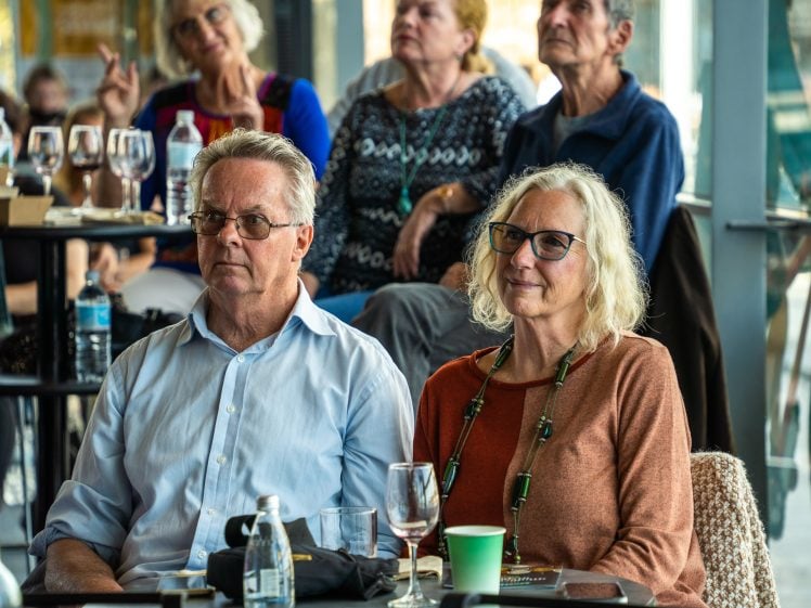 Image of An older man and woman sit together at a table with drinks, attentively watching something off-camera. Other people are seated in the background. The scene appears to be a casual gathering or event in a bright indoor space.