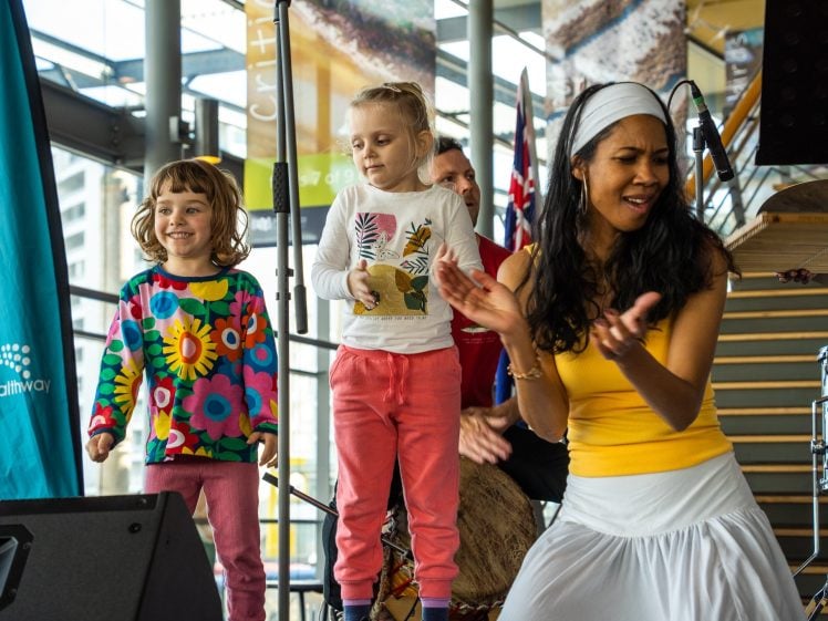 Image of A woman in a yellow top dances and claps next to two young girls in colourful clothing on a stage, with musical instruments and a drummer in the background.