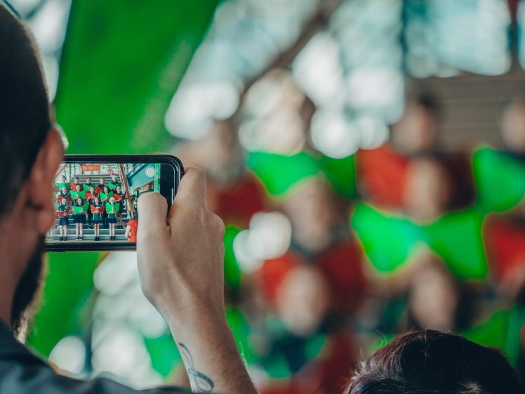 Image of A person holds up a mobile phone, taking a photo or video of a group of people dressed in red and green, who stand together in the background with a festive, blurred setting.