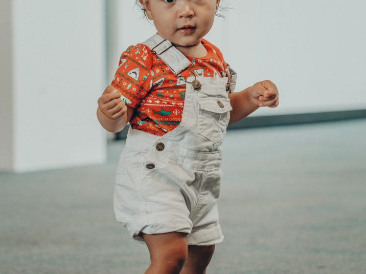 Image of A toddler wearing a red patterned shirt, white dungarees, and pink sandals stands on a blue circle indoors, looking at the camera. The background features a blurred wall with framed pictures.