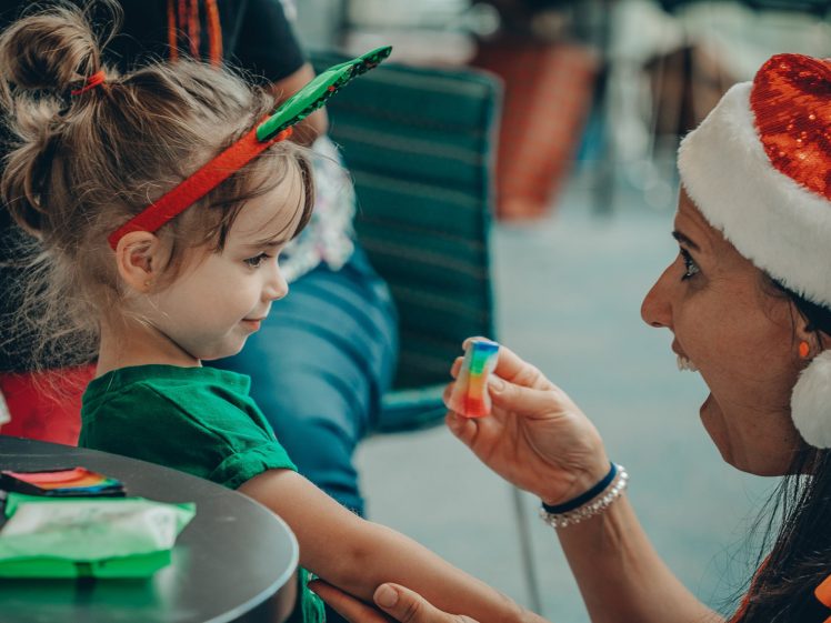 Image of A woman in a Santa hat smiles and holds face paint whilst a young girl wearing reindeer antlers and a green shirt looks on, sitting at a table with art supplies.
