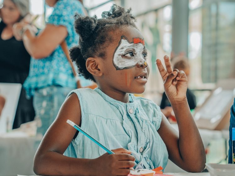 Image of A young child with face paint sits at a table, holding up two fingers in a peace sign while drawing with coloured pencils. The child wears a light blue dress and smiles, with people blurred in the background.