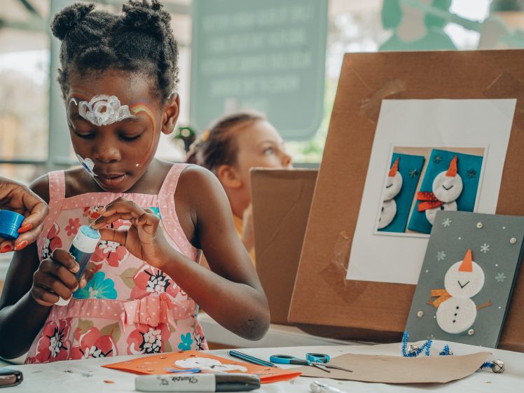 Image of A young girl in a pink floral dress with face paint crafts a snowman figure at a table. Art supplies and snowman artwork are scattered around her as she focuses on her project.
