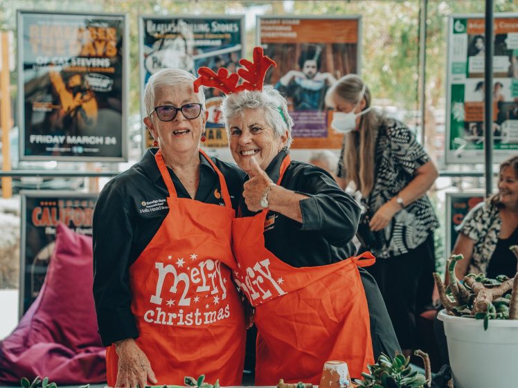 Image of Two older women wearing orange “Merry Christmas” aprons smile at the camera; one gives a thumbs-up and wears reindeer antlers. People and festive posters are visible in the background.
