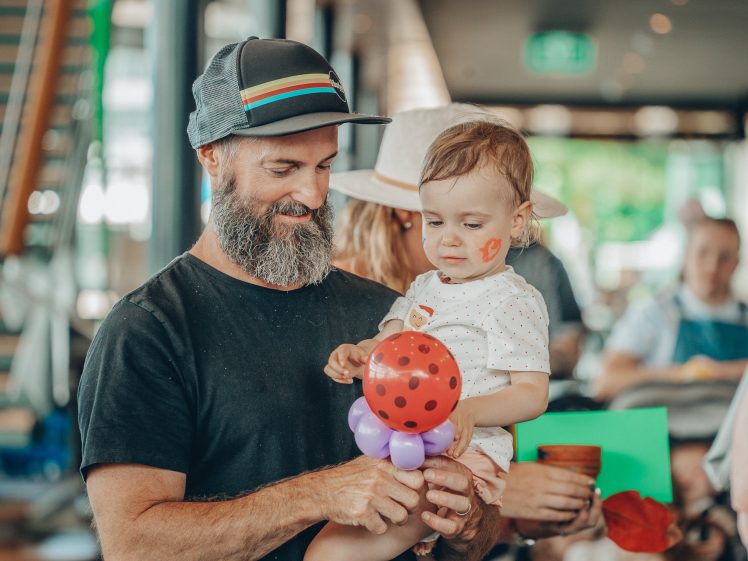 Image of A bearded man in a cap holds a child with a ladybird balloon and a ladybird face painting indoors at a lively gathering. The child wears a white outfit and looks at the balloon. Other people are blurred in the background.
