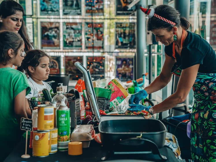 Image of A woman wearing festive accessories prepares food at a table while three children and one adult watch. The table holds cooking supplies, tinned goods, and kitchen equipment. Colourful decorations and posters are visible in the background.