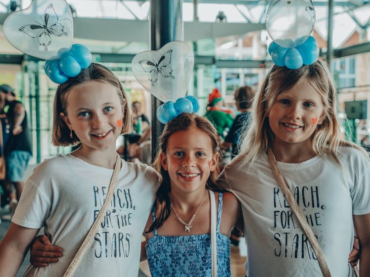 Image of Three smiling girls pose together indoors. Two wear matching shirts that say Reach for the Stars and butterfly balloon headbands. The third wears a blue dress. People and light stream through windows in the background.