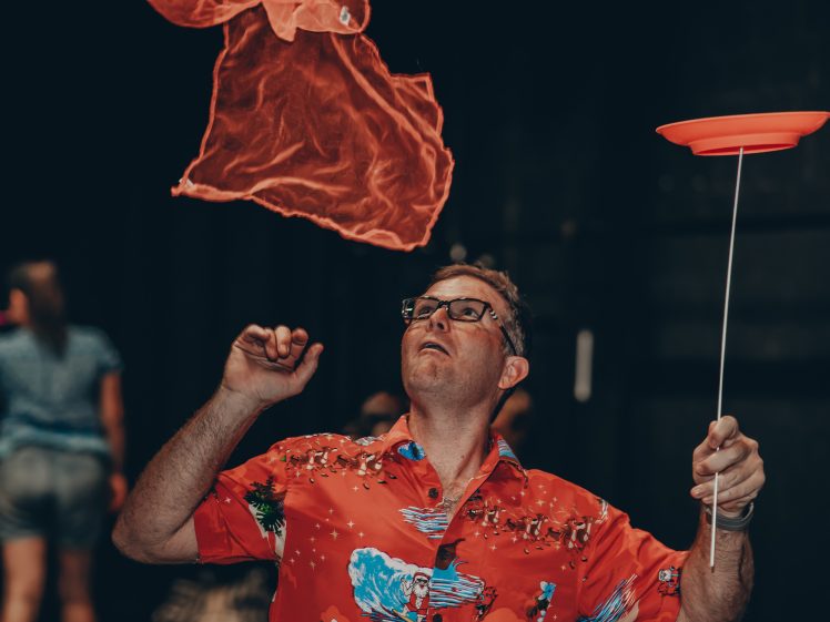 Image of A man in a bright red Hawaiian shirt balances a spinning red plate on a stick and watches an orange scarf floating above him. The background is dimly lit with blurry figures.