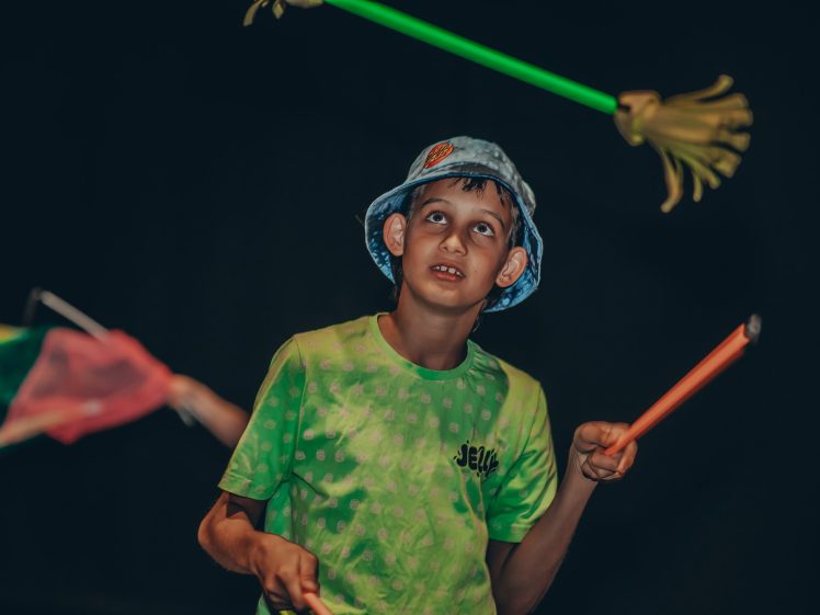 Image of A boy wearing a light blue bucket hat and a green shirt looks up while spinning juggling sticks, with colourful props blurred in the background against a dark backdrop.