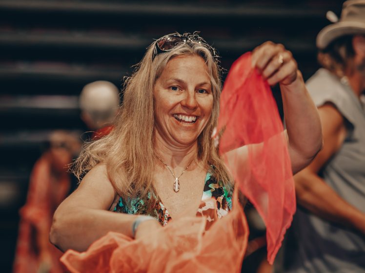 Image of A smiling woman with long blonde hair holds up a flowing, translucent orange scarf. She is wearing a floral dress and sunglasses on her head, with people and a blurred background behind her.
