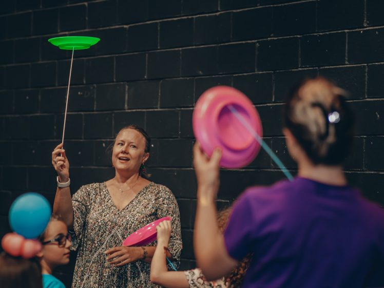 Image of A woman and children spin colourful plates on sticks in front of a dark brick wall. The woman smiles whilst participating in the activity. One child wears a blue balloon hat.