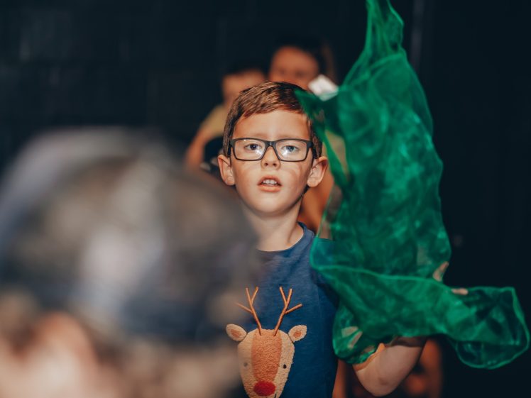 Image of A young boy wearing glasses and a blue shirt with a reindeer design holds up a green translucent fabric, with other children blurred in the background.