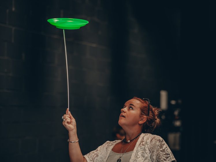 Image of A woman with auburn hair, wearing a white lace top, spins a glowing green plate on a stick whilst looking up at it against a dark background.