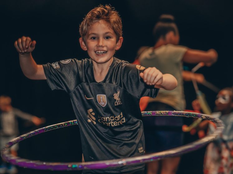 Image of A smiling boy in a dark sports kit hula hoops, with other children playing in the blurred background. The scene is lively and joyful.