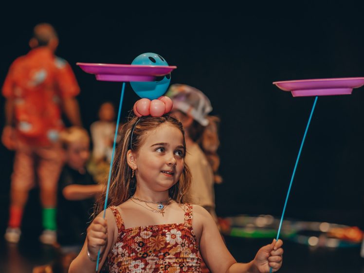 Image of A young girl wearing a flowered dress and a balloon hat balances two pink spinning plates on blue sticks, smiling and looking to the side. In the blurred background, other children and adults are visible.