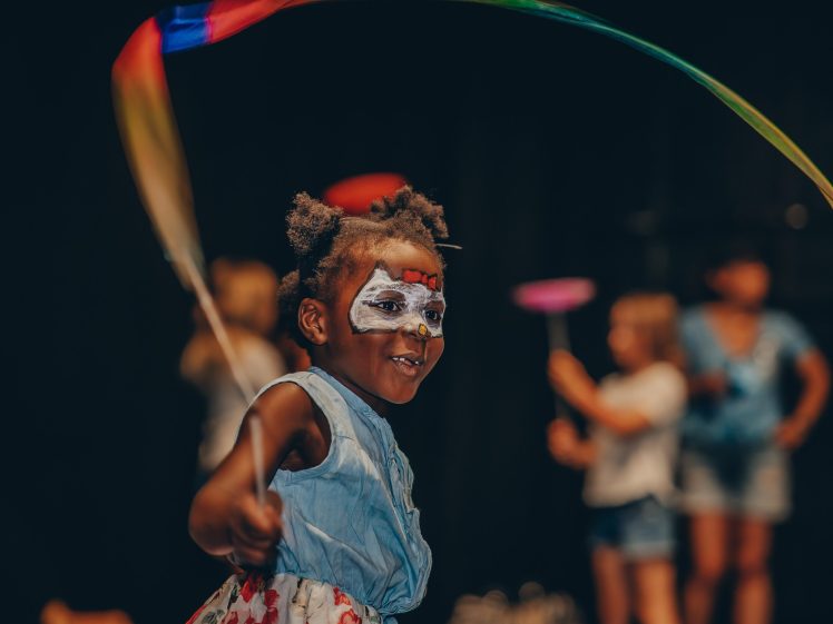 Image of A young child with face paint smiles whilst waving a colourful ribbon. She wears a light blue dress and stands in the foreground, with other children playing in the blurred background.