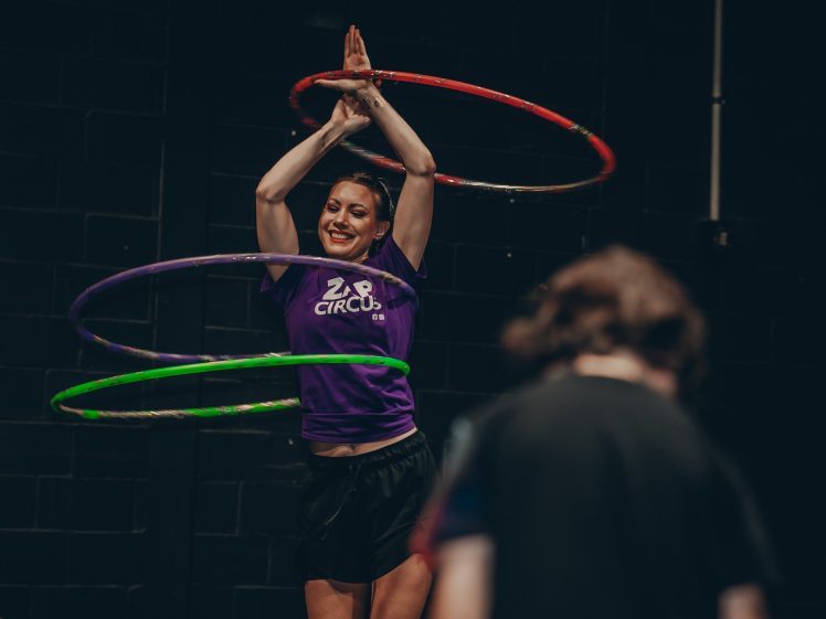 Image of A woman in a purple ZAP CIRCUS shirt spins three colourful hula hoops around her body and smiles whilst performing indoors. Another person is visible in the foreground, out of focus.