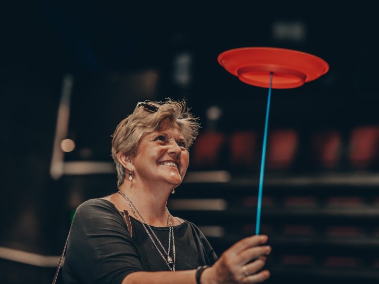 Image of A smiling woman stands indoors, spinning a red plastic plate on a blue stick. The background is blurred, showing rows of seats, suggesting she is in a theatre or auditorium.