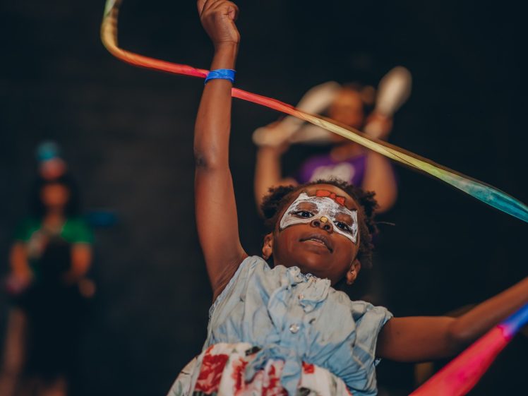 Image of A child wearing a painted mask and a blue dress joyfully waves a colourful ribbon through the air, with blurred people in the background.