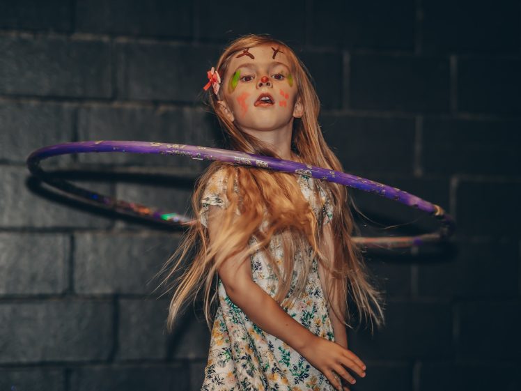 Image of A young girl with long blonde hair and face paint spins a purple hula hoop around her waist. She is wearing a floral dress and standing in front of a dark brick wall.