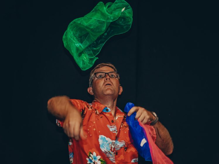 Image of A man in a bright red, floral shirt tosses colourful scarves in the air against a dark background, focussed on juggling.