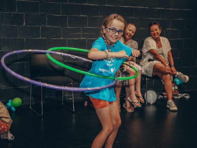 Image of A young girl with glasses smiles whilst spinning two hula hoops. She wears a blue shirt and orange shorts. Two women sit in the background, watching her and smiling. Balloons are scattered on the floor nearby.
