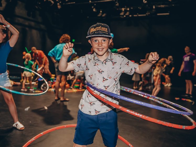 Image of A smiling boy in a patterned shirt and hat spins colourful hula hoops around his waist in a lively indoor room filled with other children playing with hula hoops.
