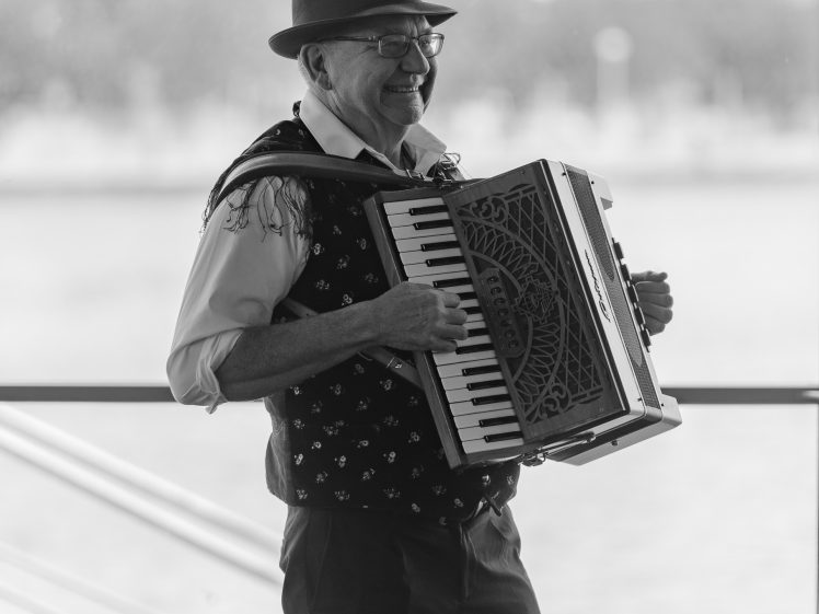 Image of An older man wearing a hat and waistcoat smiles whilst playing an accordion. The background is blurred, showing large windows, trees, and buildings, suggesting he is performing indoors near a waterfront.