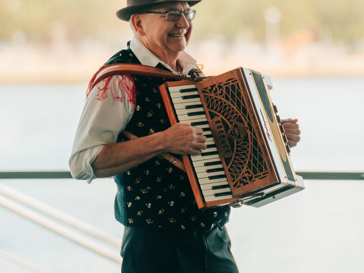 Image of An older man wearing a hat, glasses, and a patterned waistcoat smiles while playing an accordion outdoors near a waterfront.