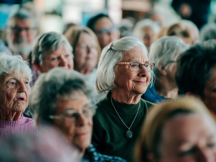Image of A group of elderly people sit closely together in an audience. Most are women, some wearing glasses. They appear engaged and attentive, with a few smiling. The background is softly focussed.