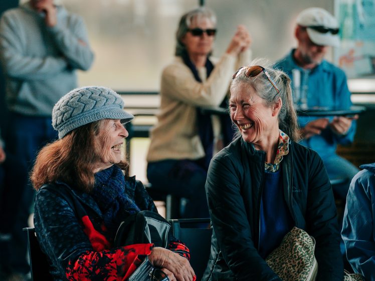 Image of Two women sit together indoors, laughing and talking, while a small group of people sit or stand in the background, some watching and some talking, in a casual, relaxed setting.