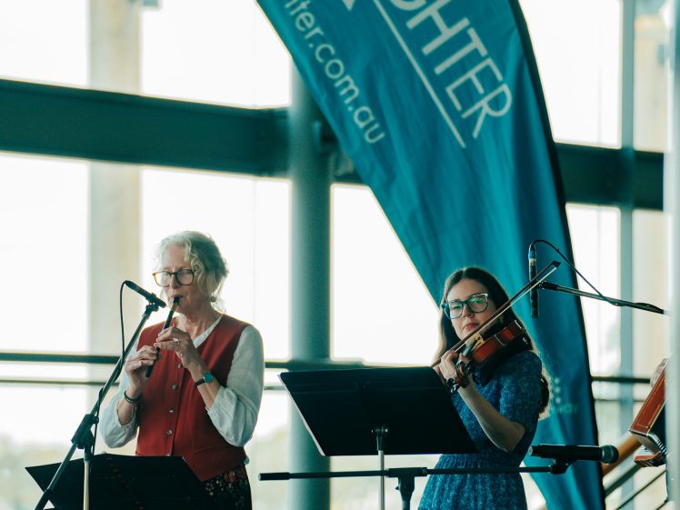 Image of Two musicians perform indoors in front of a large blue LiveLighter banner; one plays the flute and another plays the violin. Music stands and chairs are visible, with natural light streaming through windows.