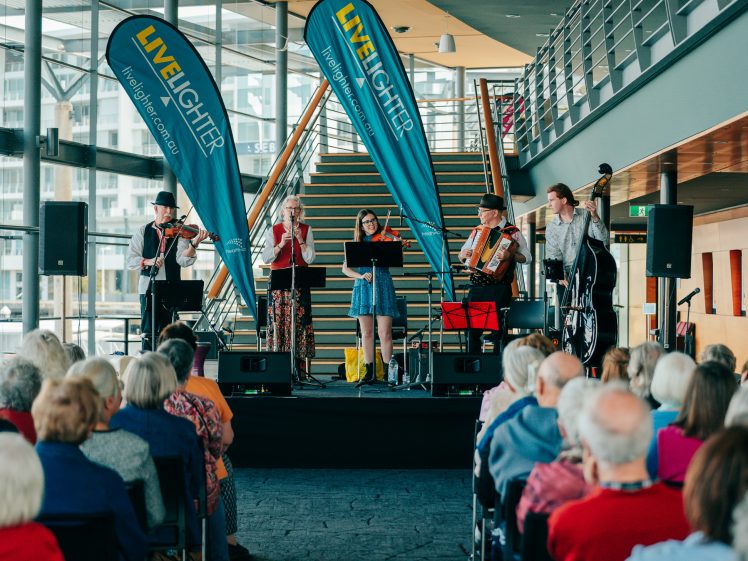 Image of A band performs on a small indoor stage for an audience. The group includes a violinist, flautist, accordionist, upright bassist, and a singer. Two LiveLighter banners are displayed behind the band.