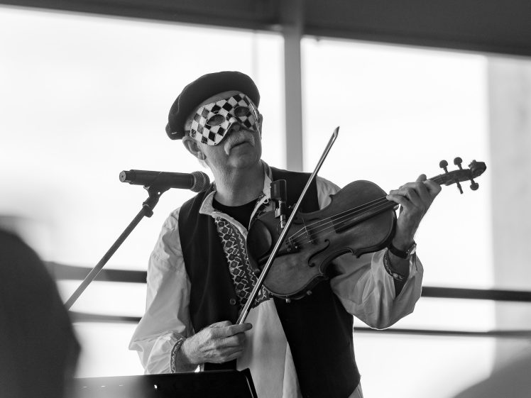 Image of A man wearing a chequered mask and beret plays the violin and sings into a microphone. He is dressed in a traditional-style shirt and waistcoat. The photo is in black and white.
