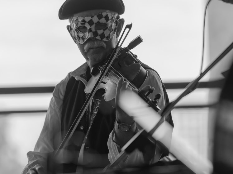 Image of A person wearing a chequered mask and beret plays the violin indoors, with musical equipment and blurred objects visible in the foreground. The image is in black and white.
