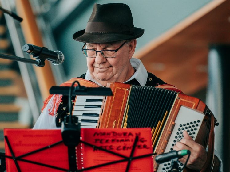 Image of An older man wearing glasses and a brown hat plays an accordion while standing behind a red music stand and a microphone. The background includes modern architectural elements and a staircase.