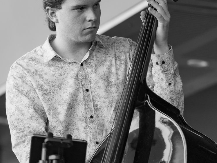 Image of A young man in a patterned shirt plays a double bass with focus. A music stand is in front of him. The image is black and white, highlighting reflections on the instrument.