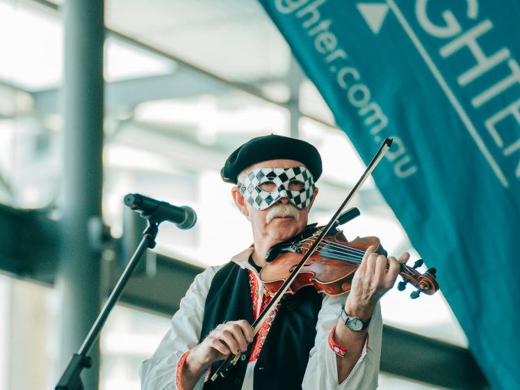 Image of A man wearing a black beret, patterned mask, and traditional clothing plays a violin on stage in front of a microphone. A large LiveLighter banner is displayed in the background.
