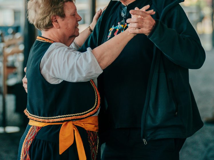 Image of An older woman and man dance together indoors. The woman wears traditional clothing with a yellow sash, whilst the man wears a dark cap and jacket. They hold hands and look at each other, appearing engaged in the dance.