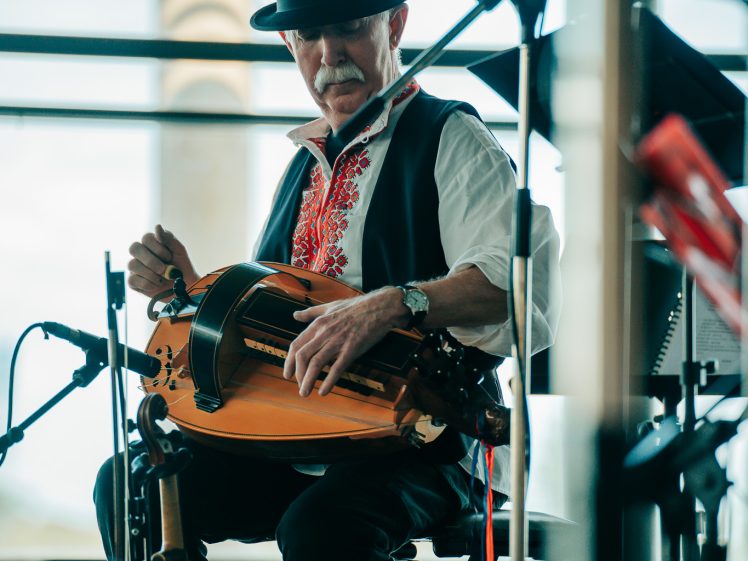 Image of A man wearing a black hat and traditional embroidered shirt plays a hurdy-gurdy whilst seated on stage, surrounded by microphones and equipment, with large windows and a blurred outdoor background.